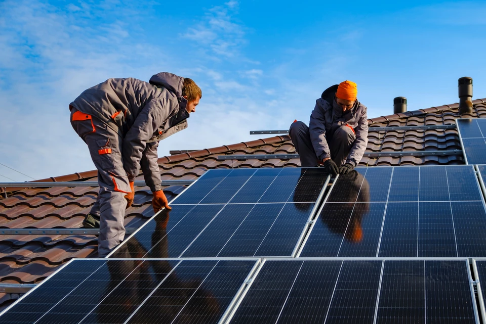Deux techniciens qualifiés en tenue professionnelle et équipement de sécurité installent une pompe à chaleur air-air sur la façade extérieure d'une maison résidentielle, par beau temps. L'installation montre le professionnalisme et la rigueur d'une intervention spécialisée en chauffage, incarnant le suivi personnalisé d'une entreprise familiale locale.