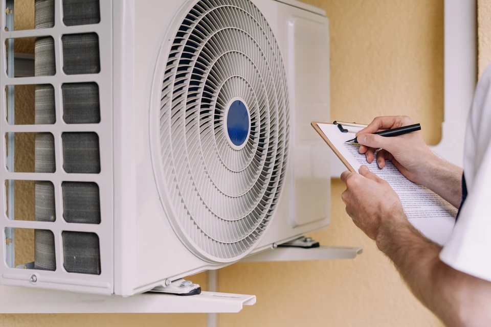 Un technicien professionnel en combinaison bleue se tenant devant une pompe à chaleur air-air montée en externe sur la façade d'une maison. Le technicien est en train de vérifier ou nettoyer les composants de l'unité avec des outils appropriés. L'image montre un bâtiment résidentiel moderne en arrière-plan, évoquant le contexte familial de Vitry-le-François. La scène doit transmettre la professionnalité, la sérénité et la confiance liée à l'entretien préventif d'un équipement de chauffage.