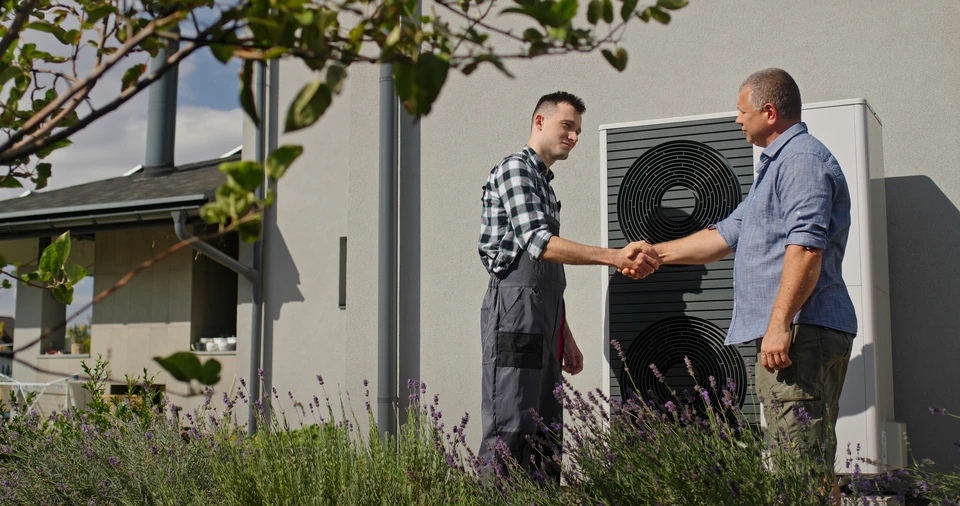 Technicien en salopette bleue installant une unité extérieure de climatisation sur mur en briques blanches, positionné sur échelle métallique